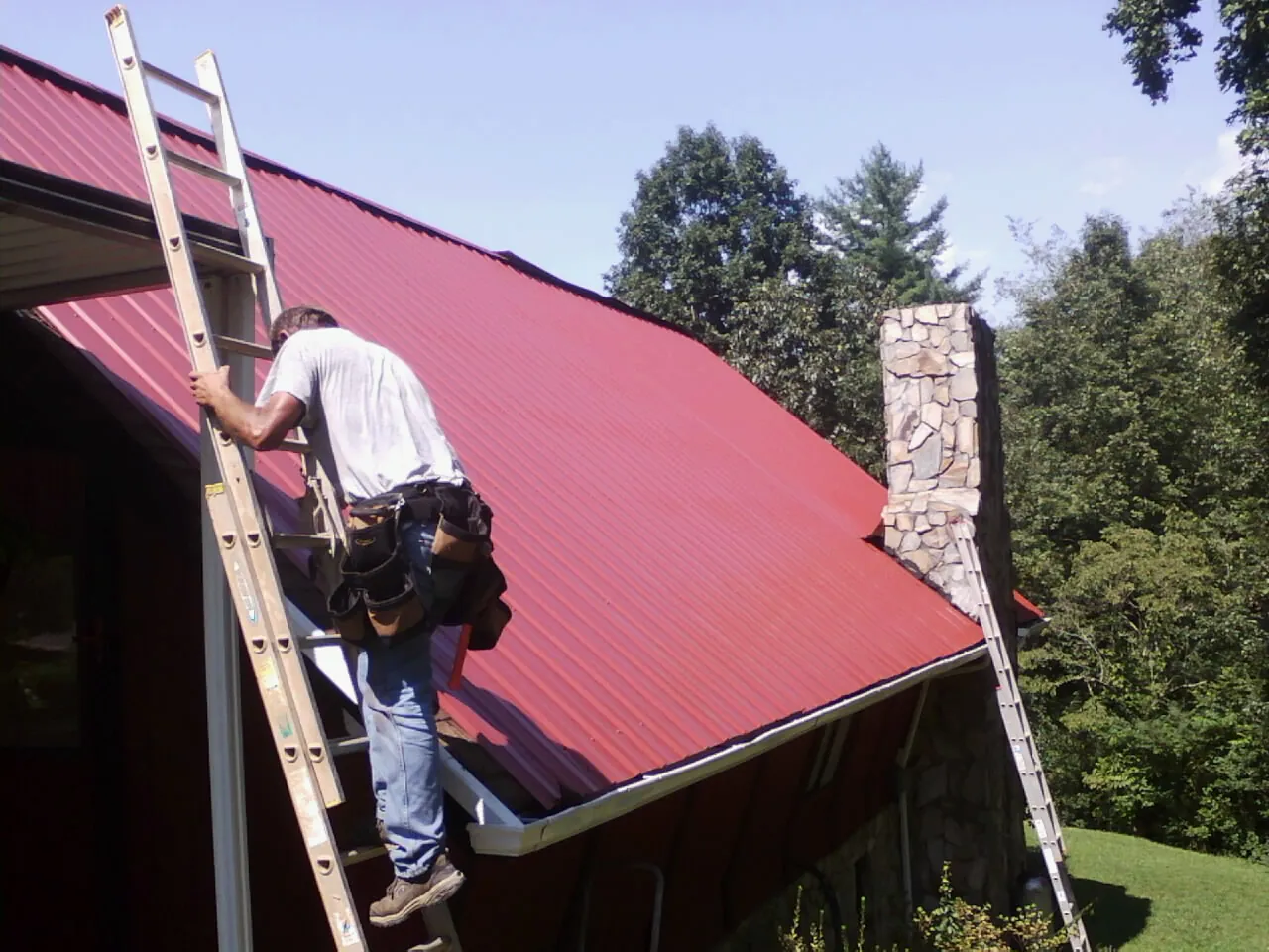 Roofer on a ladder beside a red metal roof and stone chimney — Soffit & Fascia Repair by Roofing Brothers in Thurmond